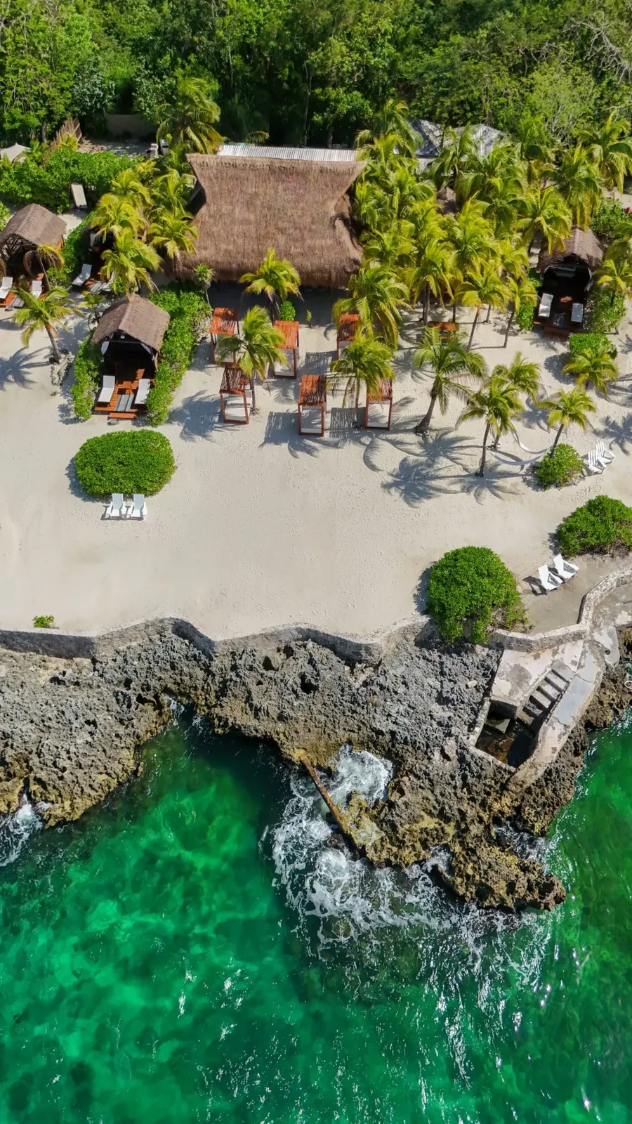 Aerial view of a Cozumel beach club with thatched cabanas, white loungers, palms, and clear turquoise water