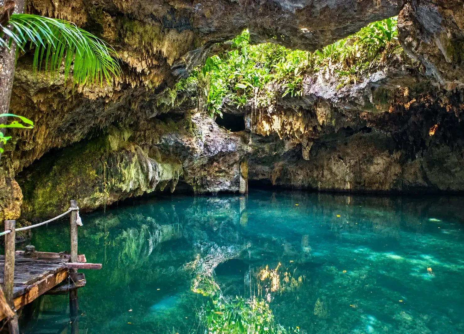 A crystal-clear turquoise cenote tucked into limestone rock, with a wooden dock at the water's edge